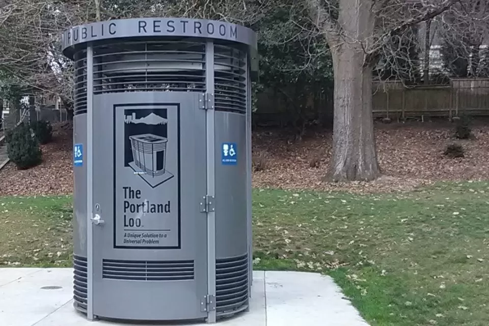 Portland Loo modular gray public bathroom booth sits on concrete slab with green lawn and tree in background in a park in Portland, Oregon